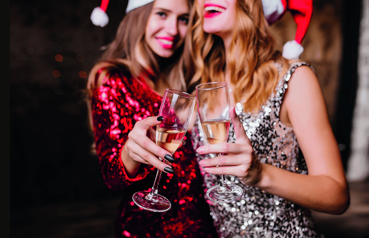 Two people in sequin dresses and Santa hats toast with champagne at a festive party venue, laughing together.