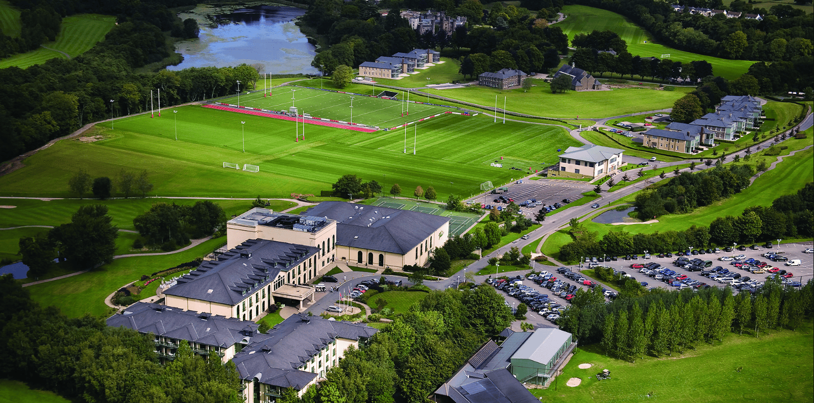 Aerial view of a sports estate complex with pitches, buildings, car park, and surrounding trees.