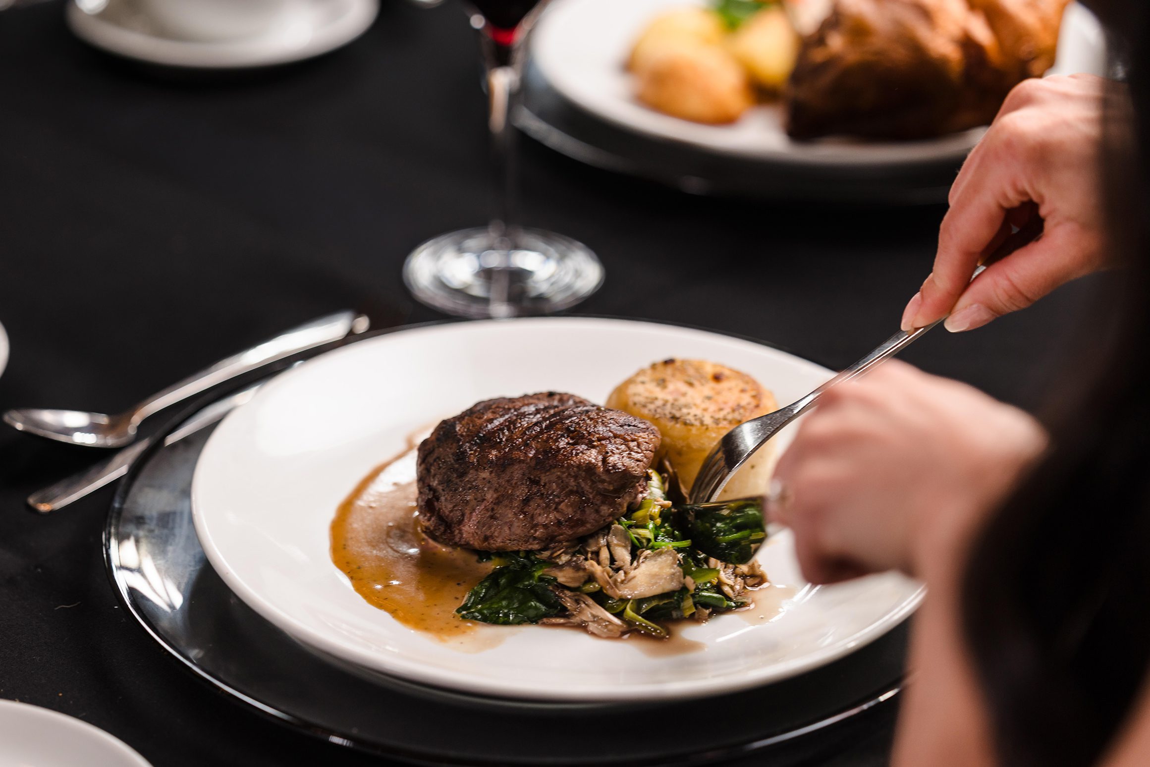 A person cuts into a steak served with greens, mushrooms, and a potato on a white plate with brown sauce.