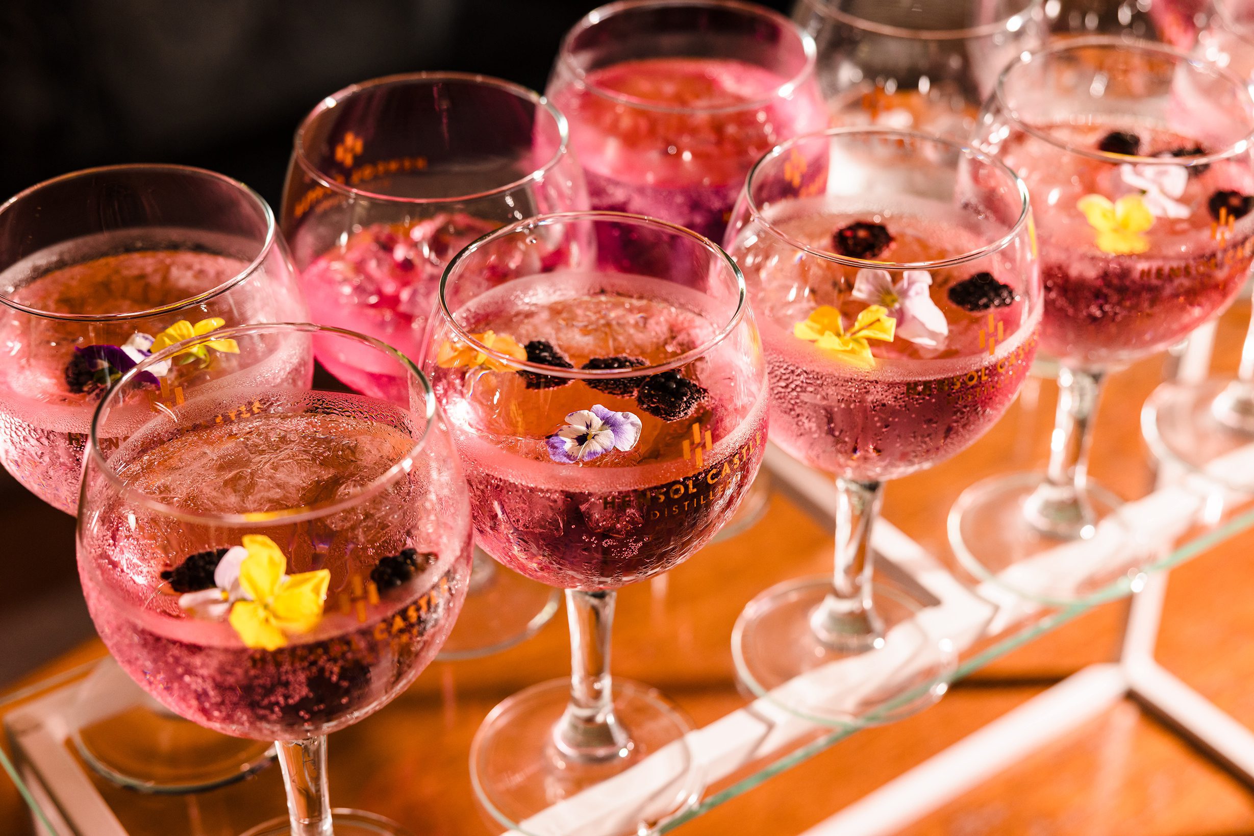 Glasses of pink cocktails with ice, berries, and edible flowers arranged on a glass tray.