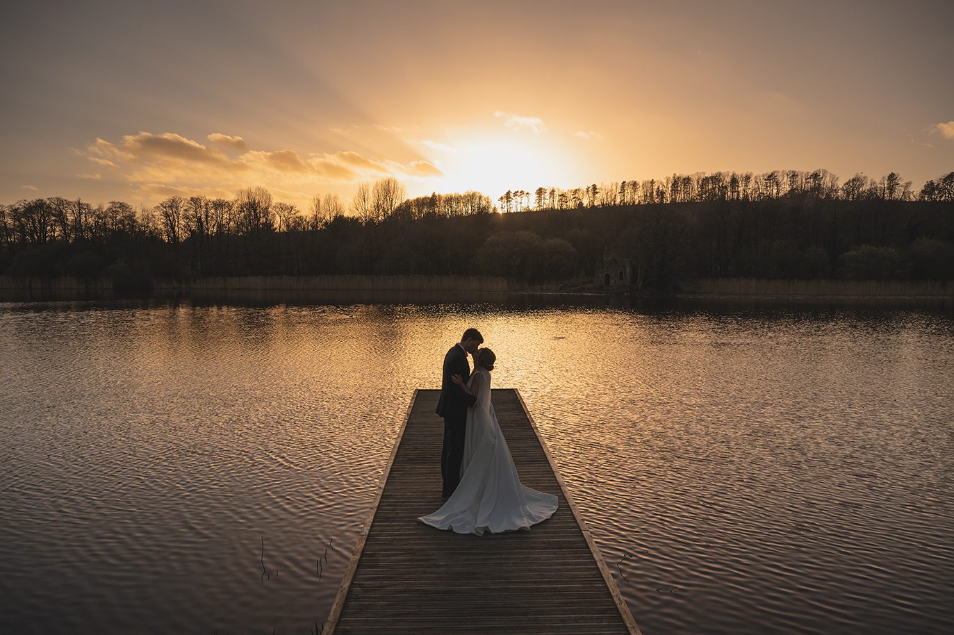 A bride and groom embrace on a jetty at sunset during their September wedding, calm water and trees behind them.