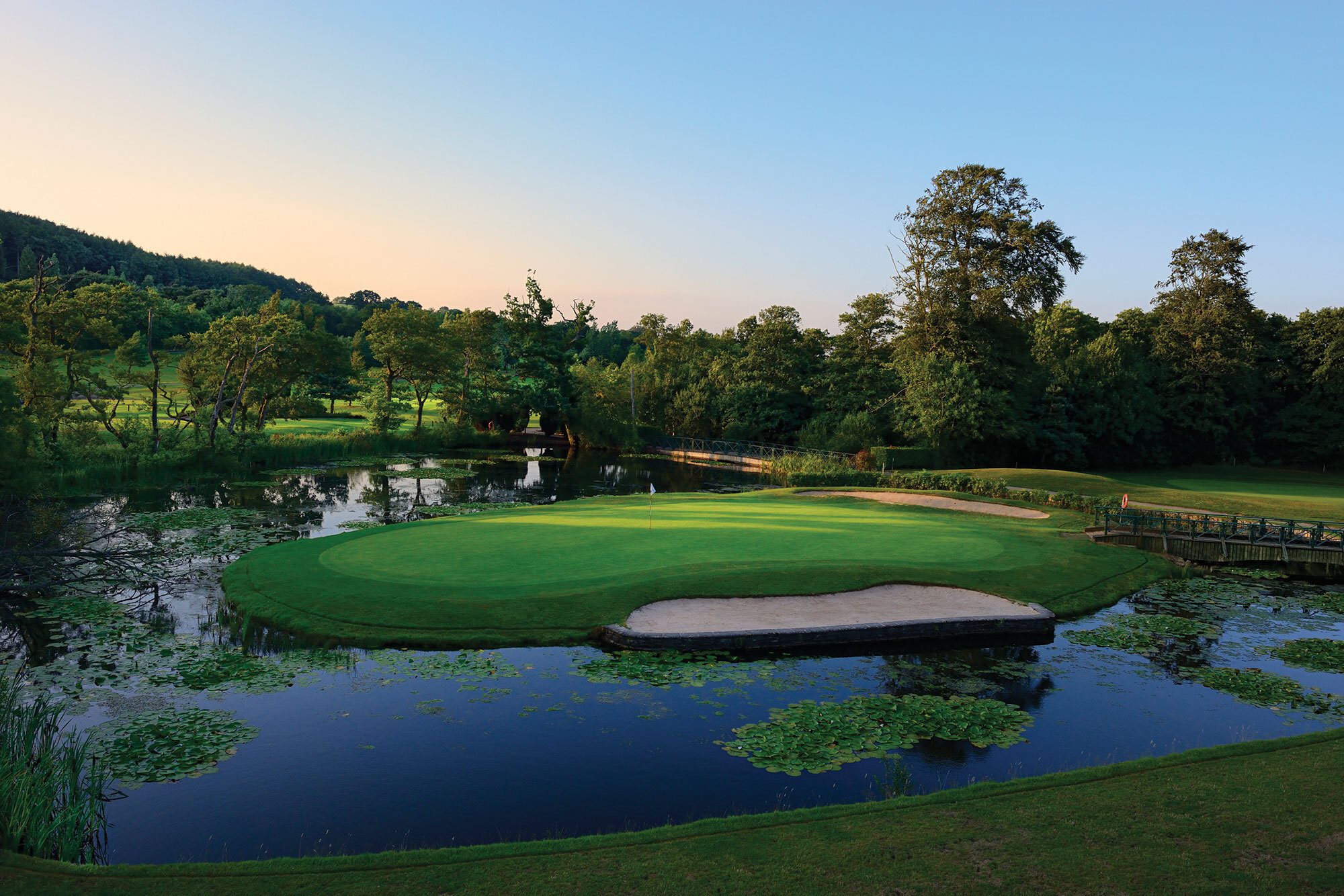 Golf green surrounded by water and trees, perfect for golf breaks Wales at sunset under a clear sky.