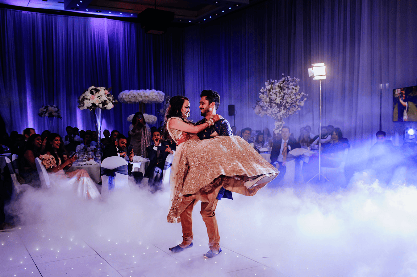 A man lifts a woman on a fog-covered dance floor at an Indian wedding, guests seated elegantly in the background.