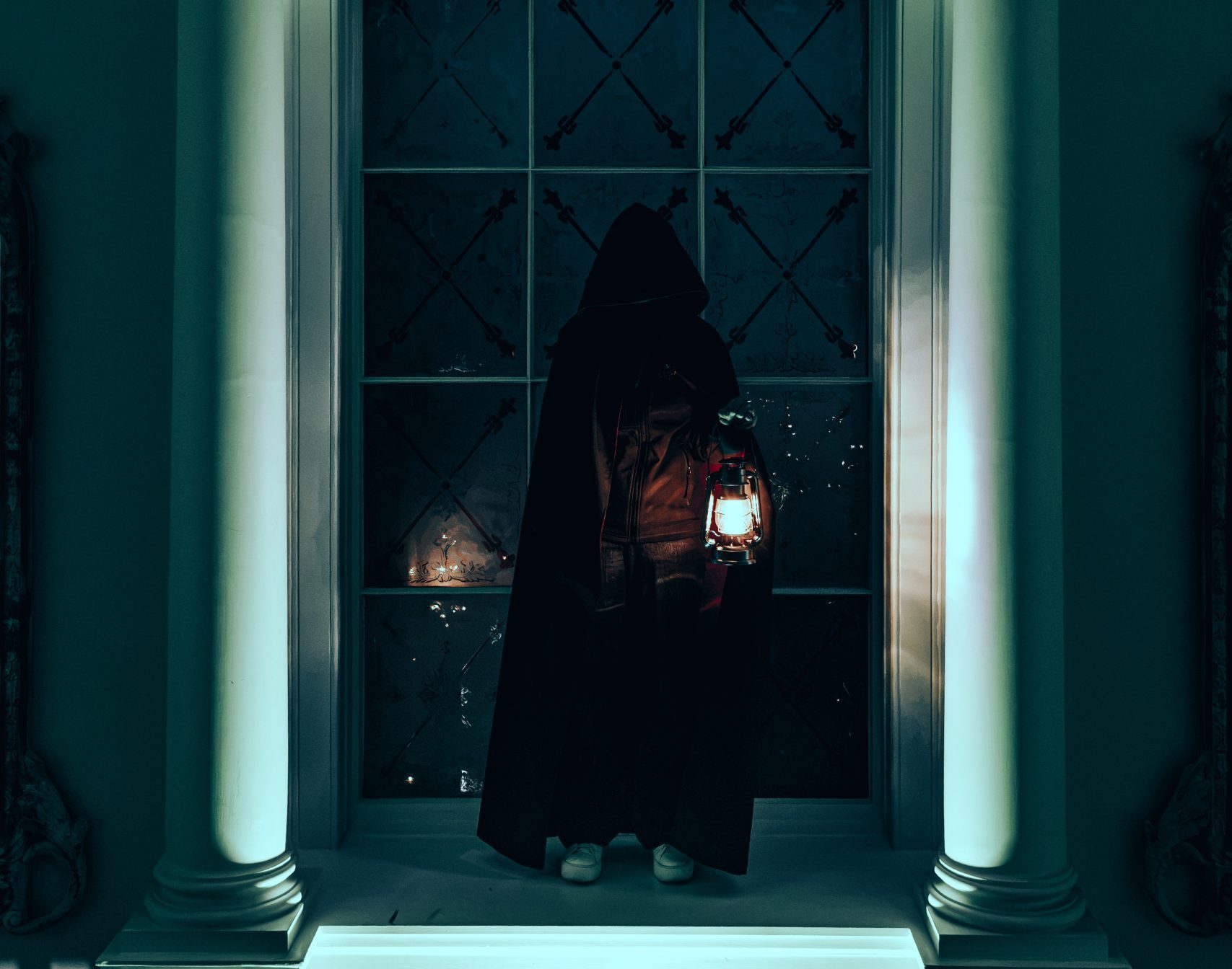A hooded figure holding a lantern stands facing a large window framed by ornate columns and an arched shell design.