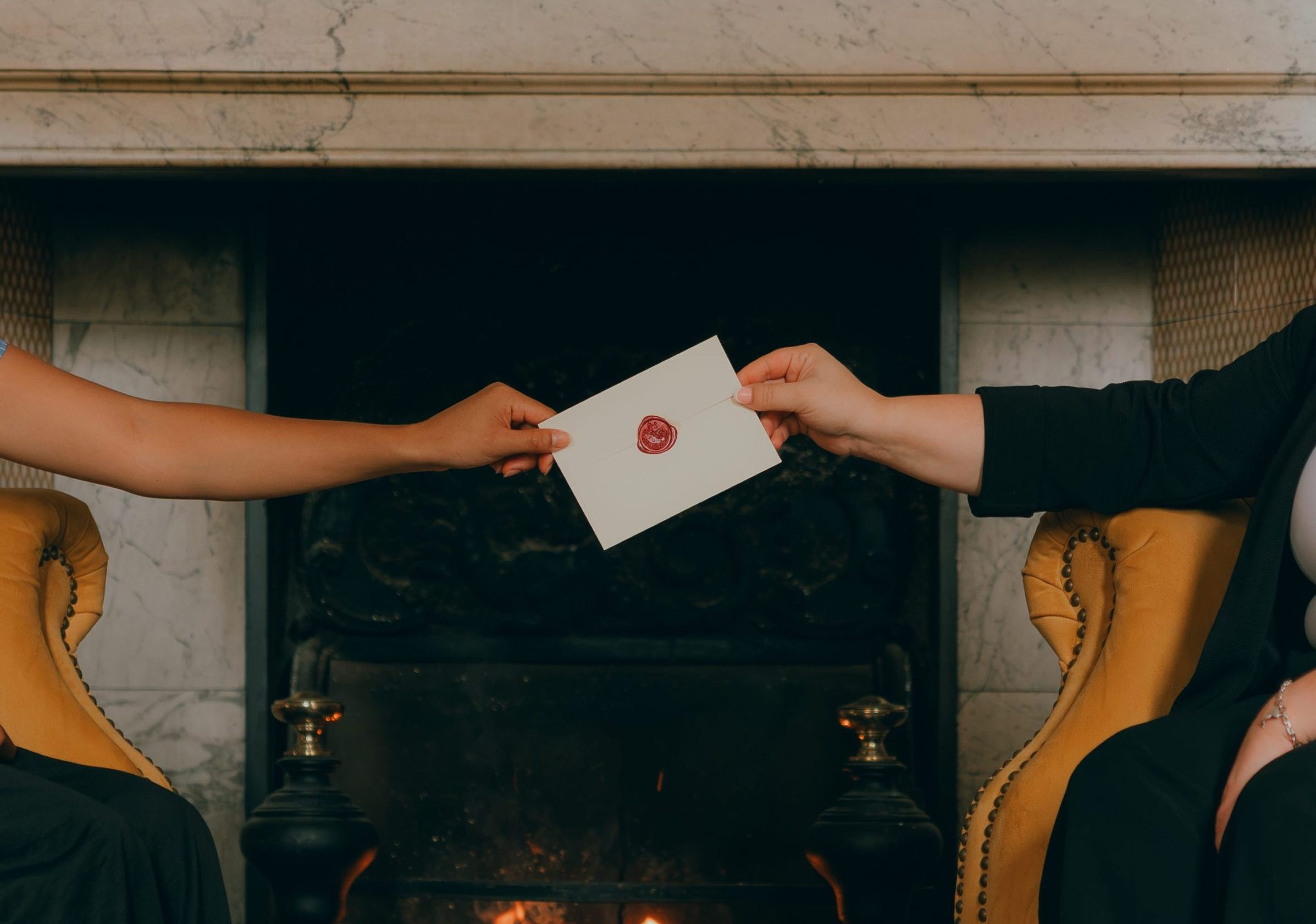Two people seated by a fireplace exchange a white envelope sealed with red wax.