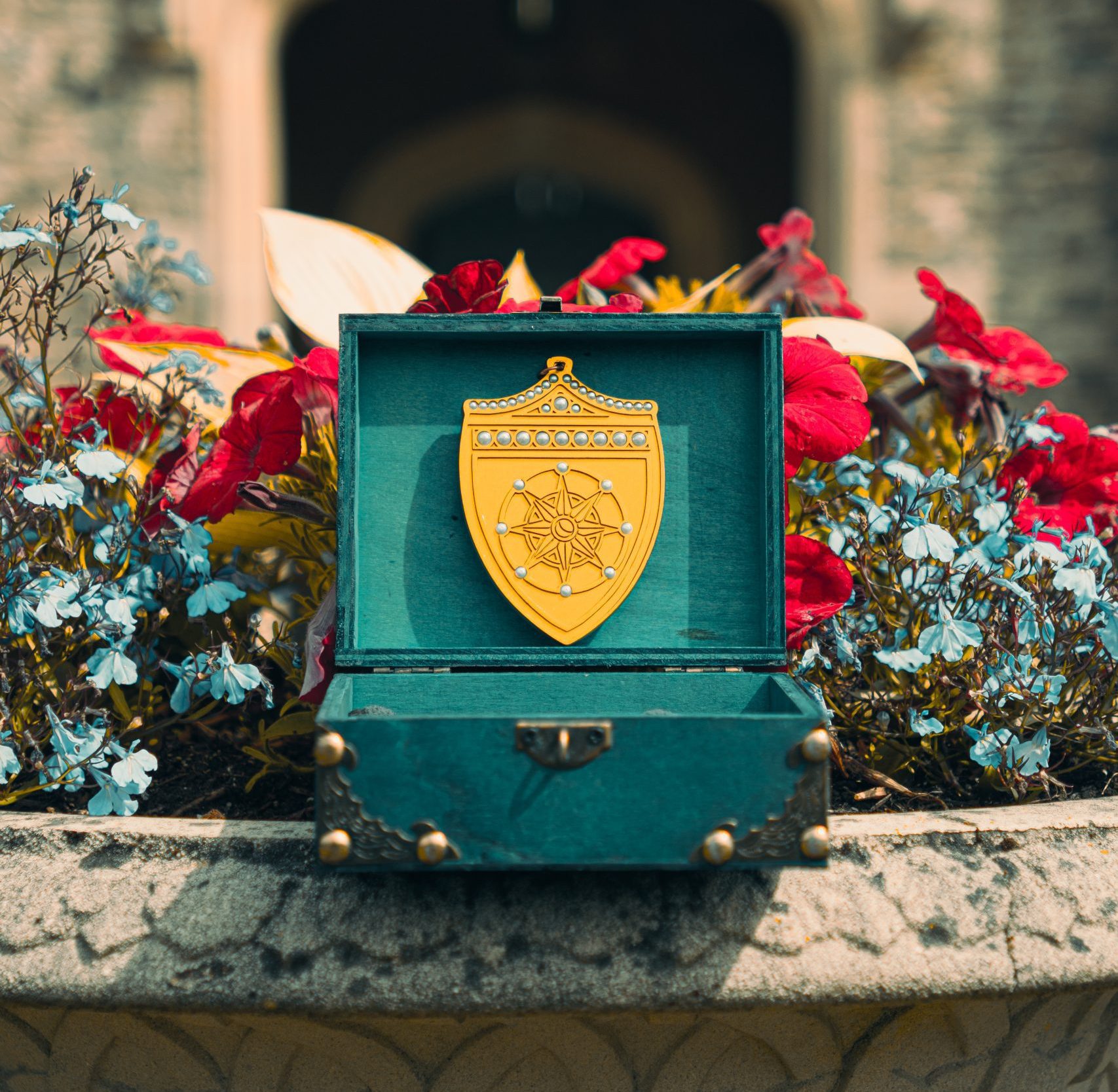 A gold shield-shaped medal in a teal box is displayed on a stone ledge with flowers and a blurred building in the background.
