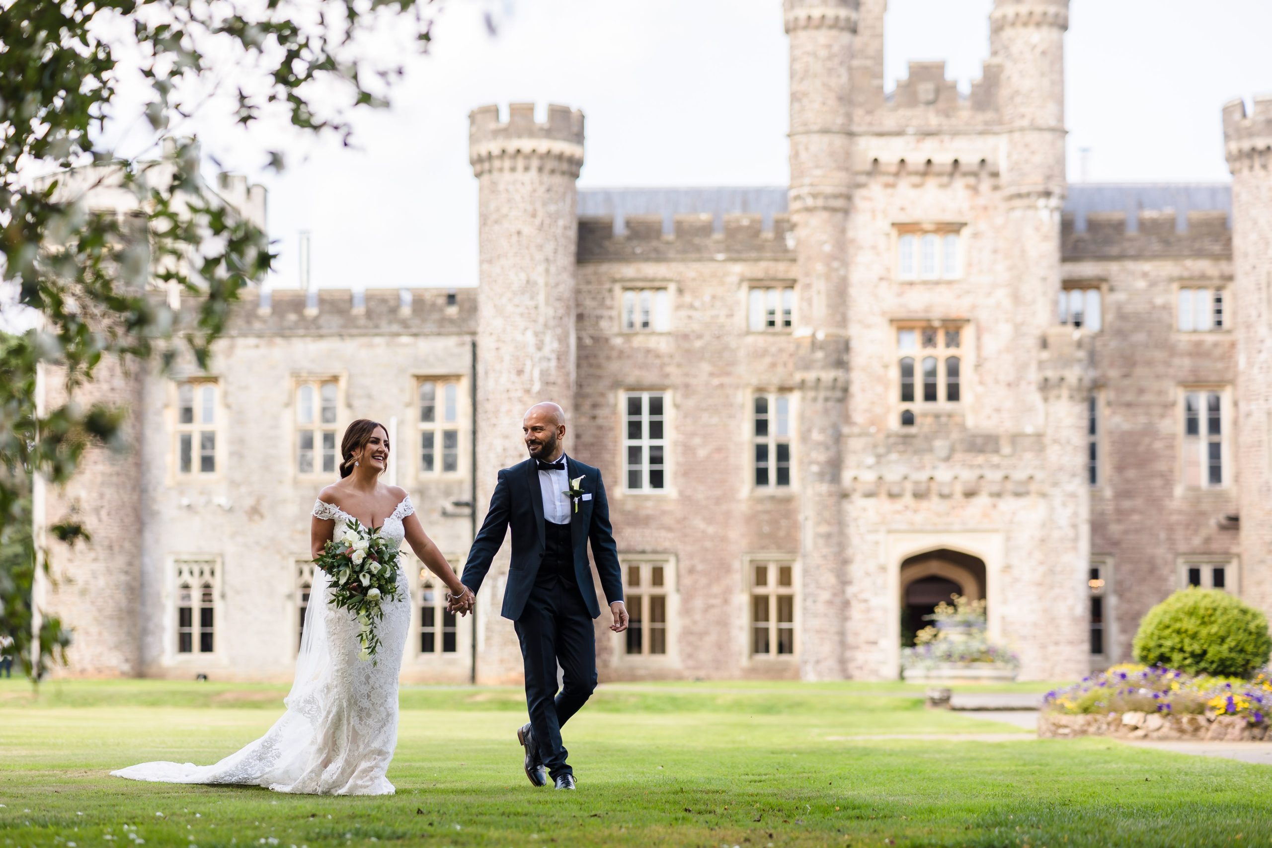 Bride and groom holding hands and walking on the lawn in front of a large stone castle at their September wedding.