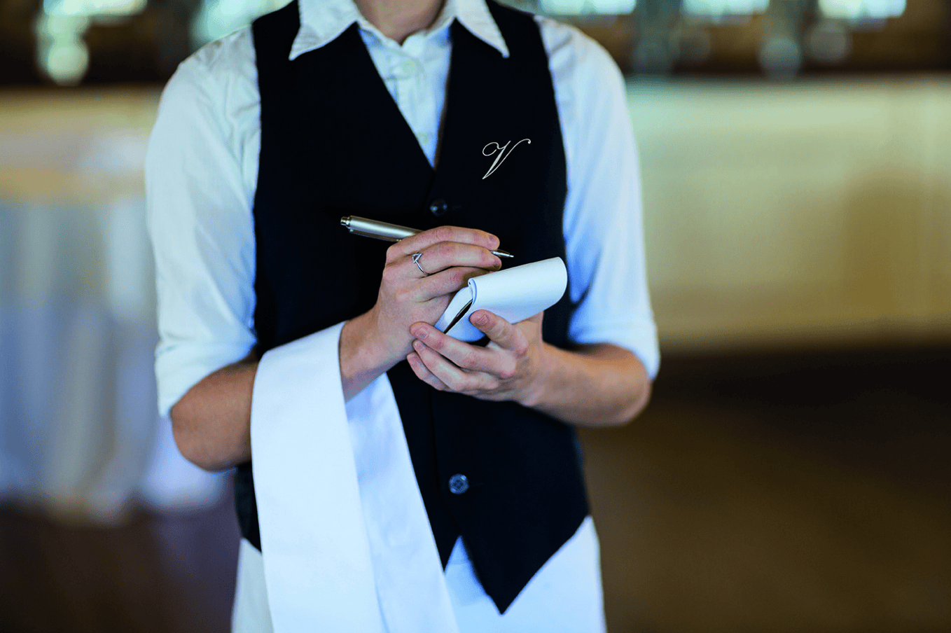 A waiter in a waistcoat, poised like estate staff, writes on a notepad with a pen, cloth folded neatly over their arm.