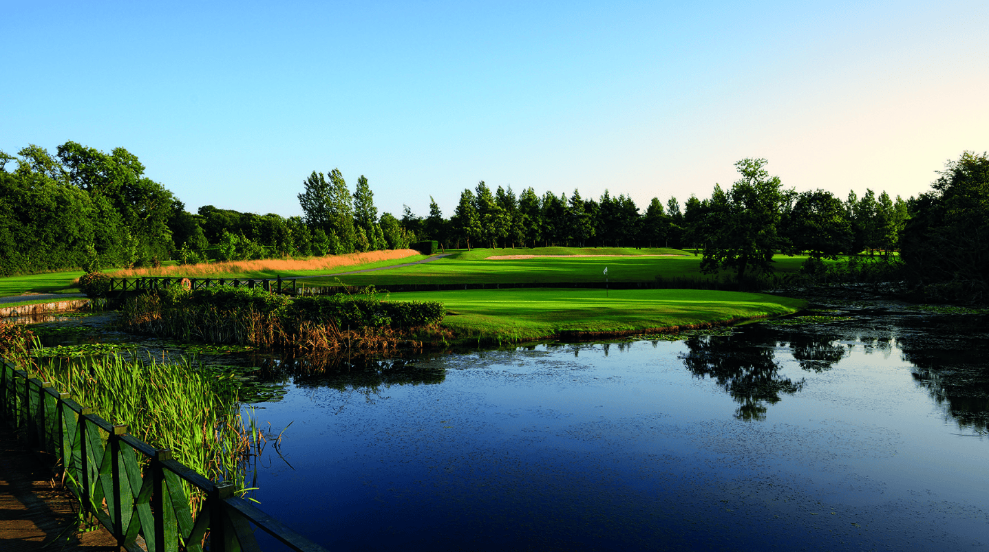 A historic destination golf course with a pond in the foreground, green fairways, and trees under a blue sky.